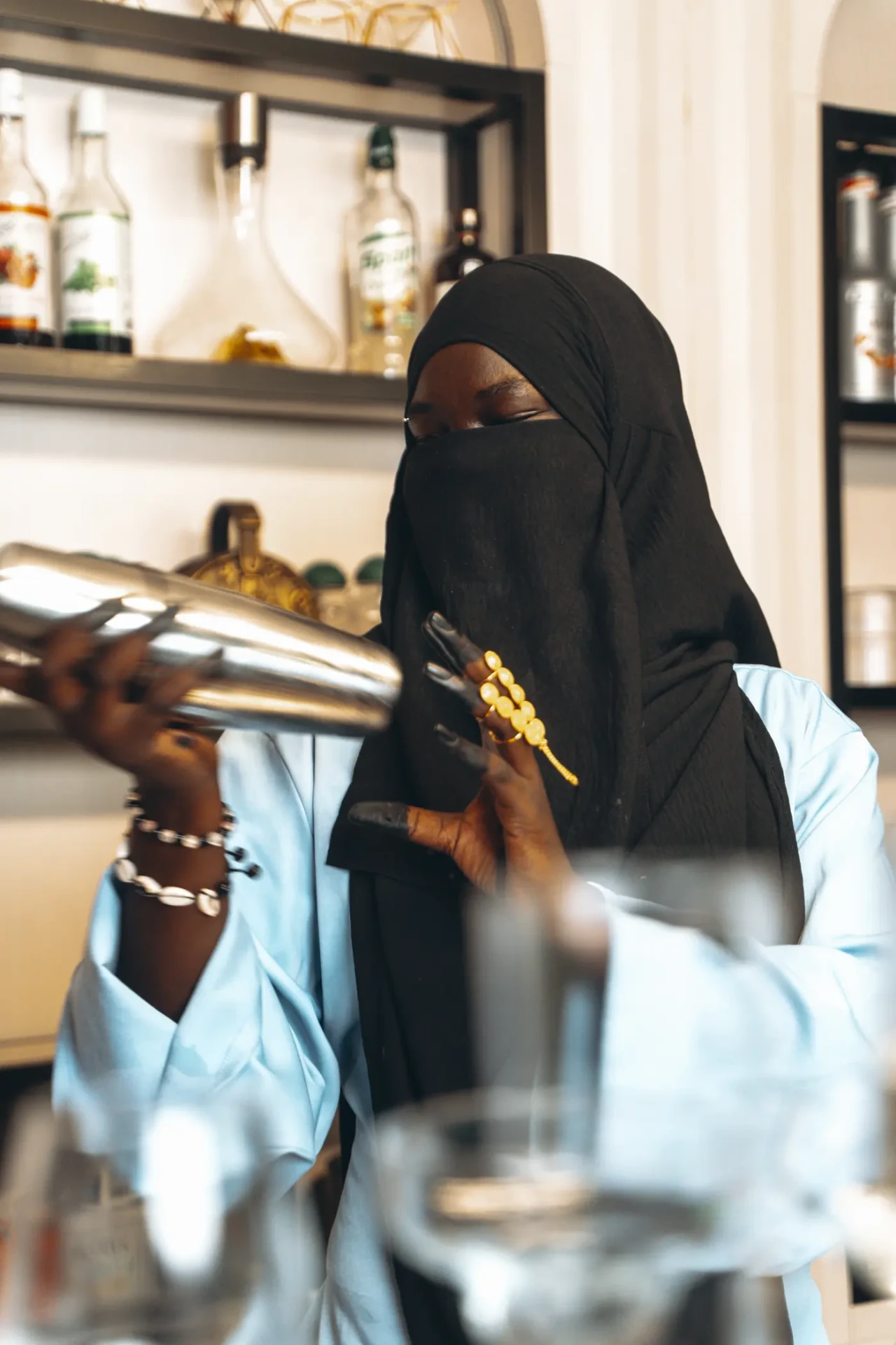 Penda Carlol secoue un shaker derrière un bar, verrerie au premier plan