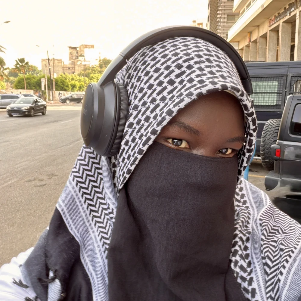 Portrait de Penda Carlol au keffieh noir et blanc et casque audio, dans une rue de Dakar